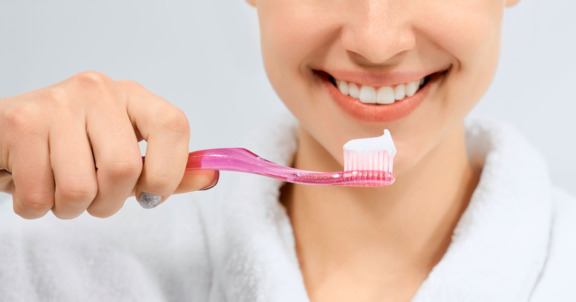 Kuvassa on pinkki hammasharja, jossa tahnaa. Harjaa pitelee hymyilevä nuori nainen, josta näkyy vain käsi, suu ja vähän valkoisen kylpytakin kaulusta.